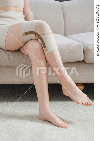 Knee support brace on a woman leg. girl in an orthosis in the interior of the house. Orthopedic anatomic braces for knee fixation, injuries and pain. Knee Joint Bandage Sleeve. Elastic Sports. Knee support brace on a woman leg. girl in an orthosis in the interior of the house. Orthopedic anatomic braces for knee fixation, injuries and pain. Knee Joint Bandage Sleeve. Elastic Sports. 92019071