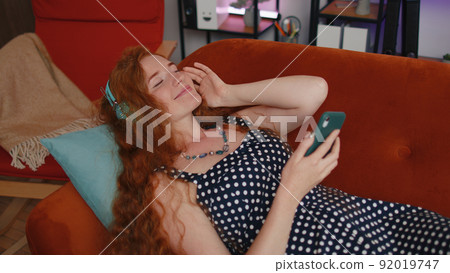 Happy freckles girl in wireless headphones relaxing lying on sofa at home listening favorite music Happy freckles girl in wireless headphones relaxing lying on sofa at home listening favorite music 92019747