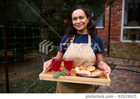 Hispanic mature woman smiles looking at camera, holding a tray of homemade pies and refreshing compote in drinking glass Hispanic mature woman smiles looking at camera, holding a tray of homemade pies and refreshing compote in drinking glass 92019925