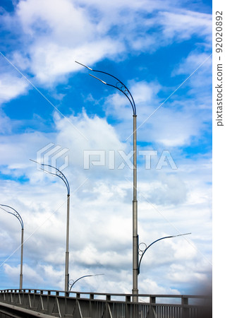 Modern LED lamp posts in a row against a blue sky with white clouds vertical wallpaper. Road lighting, electric streetlights abstract background. City illumination in daylight. Light off. Industry. 92020892