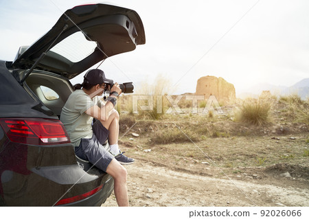 asian photographer sitting at the edge of trunk of a car taking picture of a deserted castle in a historical site 92026066