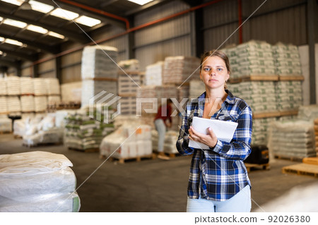 Attentive female manager keeps records of bulk construction materials in warehouse, writing down the data on paper Attentive female manager keeps records of bulk construction materials in warehouse, writing down the data on paper 92026380
