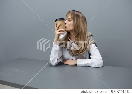 Cute happy young blonde student lady freelancer in warm sweater kiss takeaway coffee sitting at workplace in gray modern home office. Coffee break Lover Concept. Copy space. Cool offer Cute happy young blonde student lady freelancer in warm sweater kiss takeaway coffee sitting at workplace in gray modern home office. Coffee break Lover Concept. Copy space. Cool offer 92027468
