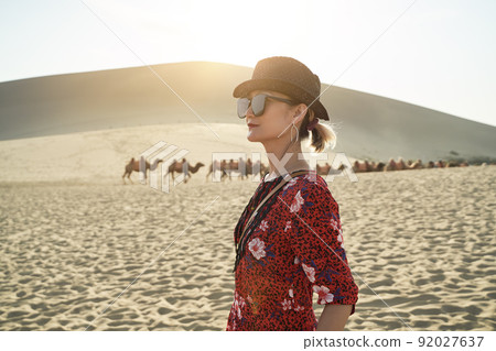 asian woman in red dress looking at view in desert with caravan of camels and huge sand dunes in background 92027637