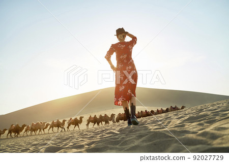 rear view of asian woman in red dress standing in desert looking at view with caravan of camels and huge sand dune in background rear view of asian woman in red dress standing in desert looking at view with caravan of camels and huge sand dune in background 92027729