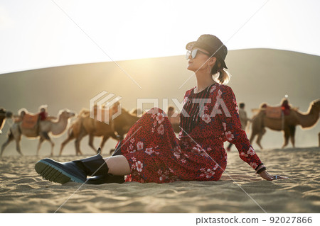 asian woman in red dress sitting in desert looking at view with caravan of camels and huge sand dune in background asian woman in red dress sitting in desert looking at view with caravan of camels and huge sand dune in background 92027866