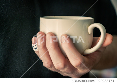 A man is holding a ceramic cup in his hand. The first phalanx of his little finger is wrapped in white ribbon. A smiling face is painted on the bandage. Positive emotion concept. Selective Focus. 92028573