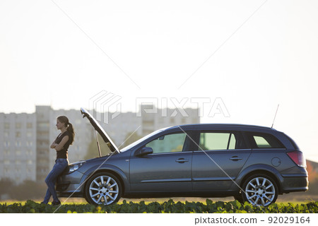 Young attractive woman at car with open hood in green meadow on blurred apartment building and clear bright sky copy space background. Transportation, vehicles problems and breakdowns concept. 92029164
