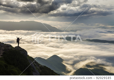 Wide mountain panorama. Small silhouette of tourist with backpack on rocky mountain slope with raised hands over valley covered with white puffy clouds. Beauty of nature, tourism and traveling concept 92029185