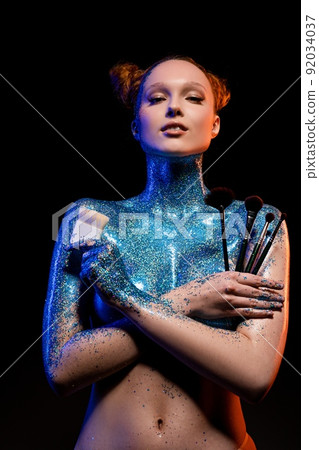 Alluring lady with glossy body art standing in studio with makeup brushes 92034037