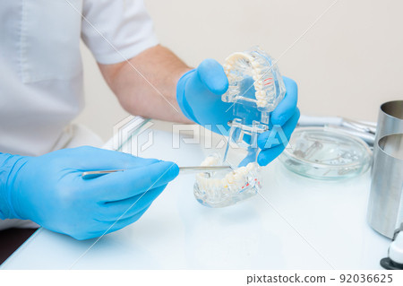 hands of a dentist in blue gloves close-up. dentist with tweezers shows defects on the acrylic model of the jaw. hands of a dentist in blue gloves close-up. dentist with tweezers shows defects on the acrylic model of the jaw. 92036625