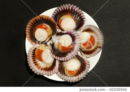 A white plate of big raw fresh scallops with orange caviar and shells on a dark background.  92036940