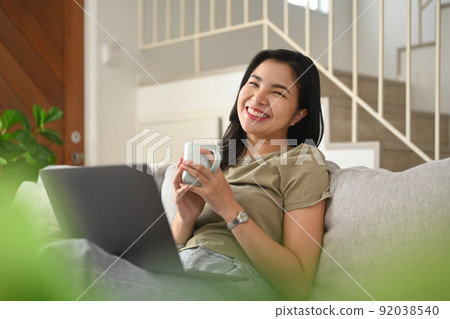 Cheerful asian woman holding coffee cup in her hands and using laptop on coat at home Cheerful asian woman holding coffee cup in her hands and using laptop on coat at home 92038540