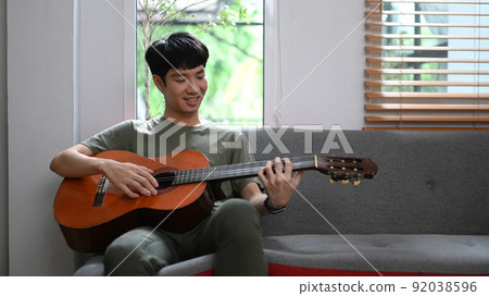 Happy asian man playing acoustic guitar on couch, enjoying carefree time at home 92038596