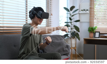 Exited man playing video game or watching movie in virtual reality headset, sitting on couch in cozy home 92038597