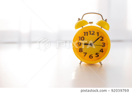 A yellow alarm clock stands on a white table by the window with white curtains A yellow alarm clock stands on a white table by the window with white curtains 92039769