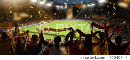 Back view of football, soccer fans cheering their team with colorful scarfs at crowded stadium at evening time. Concept of sport, support, competition. Out of focus effect 92040544