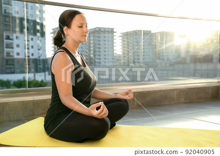 Young woman practices yoga in lotus position sitting on fitness mat on balcony of her house in rays of sun. Concept of healthy and harmonious life 92040905
