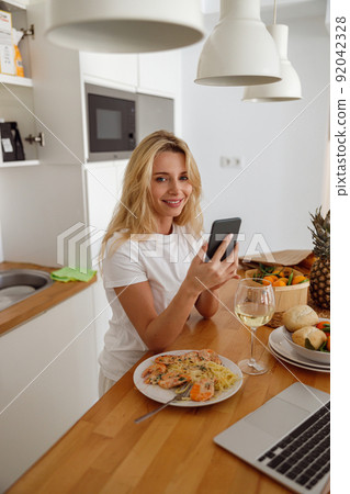 Blonde pretty woman tapping and texting on cellphone at laptop in kitchen. Vertical. 92042328