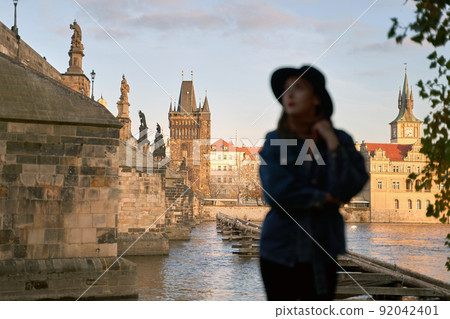Prague Silhouette. Stylish beautiful young woman wearing black hat with Charles Bridge on background. Elegant retro lady fine art portrait. Focus on background. 92042401