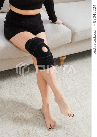 Black knee support brace on a woman leg. girl in an orthosis in the interior of the house. Orthopedic anatomic braces for knee fixation, injuries and pain. Knee Joint Bandage Sleeve. Elastic Sports. 92043224