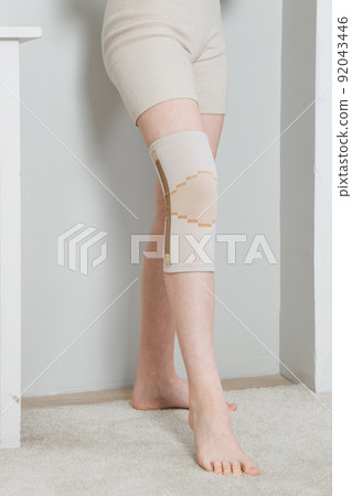 Knee support brace on a woman leg. girl in an orthosis in the interior of the house. Orthopedic anatomic braces for knee fixation, injuries and pain. Knee Joint Bandage Sleeve. Elastic Sports. 92043446