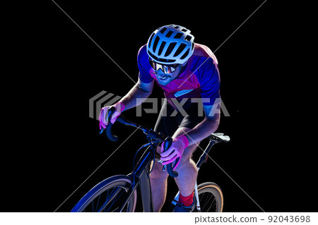 Portrait of man, professional cyclist training, riding isolated over black studio background in neon light. Outrunning 92043698