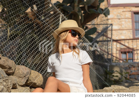 A young girl in a hat and sunglasses sits on a stone staircase. 92044085