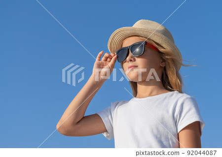A young girl in a hat and sunglasses adjusts her glasses with one hand. 92044087