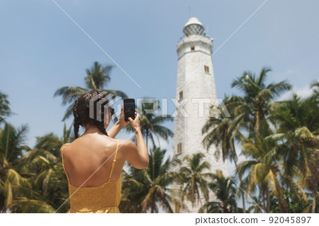 Woman traveler in dress in front of famous landmark of Sri Lanka country taking photo on smartphone of Dondra lighthouse 92045897