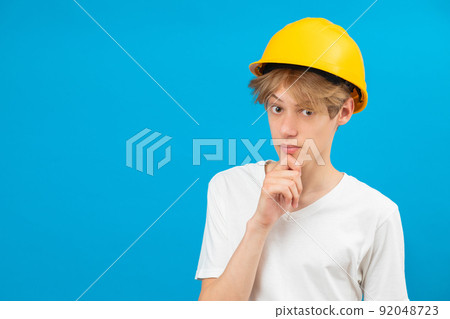 A teenage boy in a yellow helmet is looking at the camera, standing in a studio on a blue background A teenage boy in a yellow helmet is looking at the camera, standing in a studio on a blue background 92048723