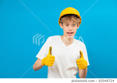Happy teen in white t-shirt and gloves in his hand with yellow helmet standing in studio on blue background. A future architect isolated on a blue background Happy teen in white t-shirt and gloves in his hand with yellow helmet standing in studio on blue background. A future architect isolated on a blue background 92048724
