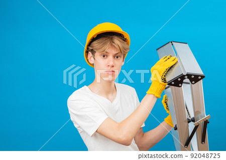 Happy teen in white t-shirt and gloves with yellow helmet in head, holding ladder in hands standing in studio on blue background. A future architect isolated on a blue background Happy teen in white t-shirt and gloves with yellow helmet in head, holding ladder in hands standing in studio on blue background. A future architect isolated on a blue background 92048725