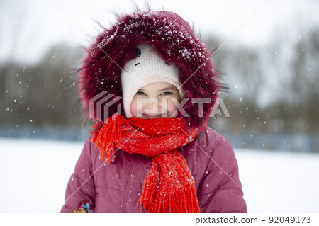 Child in winter. A little girl in a warm hat and hood looks at the camera and smiles. Child in winter. A little girl in a warm hat and hood looks at the camera and smiles. 92049173