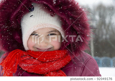 Child in winter. A little girl in a warm hat and hood looks at the camera and smiles. 92049176
