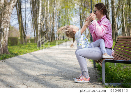 A mother cradles her daughter up and down while sitting on a bench in a city park. 92049543