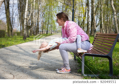 A mother and daughter play gymnastics while sitting on a bench in a city park. 92049551