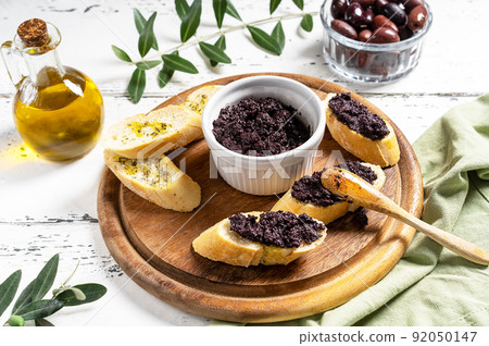 Olive pate in bowl and spreader on bread slices on wooden cutting board.  92050147
