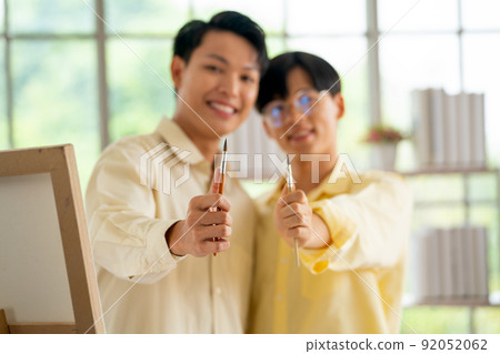 Main focus on paintbrush hold by two gay or lgbt man stand in living room with painting pallet Main focus on paintbrush hold by two gay or lgbt man stand in living room with painting pallet 92052062
