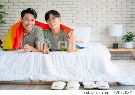 Couple gay or lgbt man use rainbow flag to cover and lie on white bed also look at camera with smile Couple gay or lgbt man use rainbow flag to cover and lie on white bed also look at camera with smile 92052087