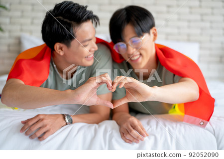 Couple gay or lgbt man with rainbow flag covering use hand to show heart symbol and they lie on bed 92052090