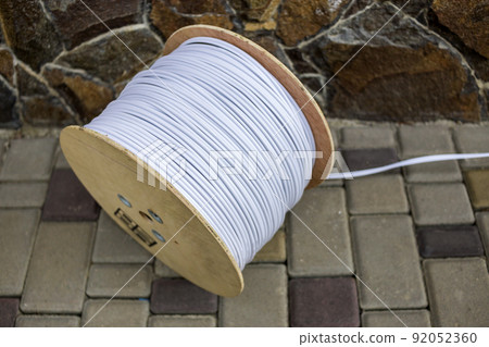 Big roll of white industrial electrical cable on large wooden reel outdoors on sunny pavement on stone fence background. Professional construction site cable, reliable wiring and durability. 92052360