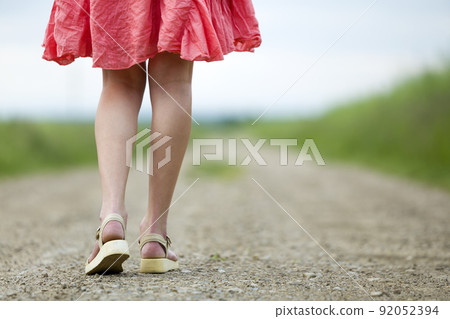 Back view of young woman in red dress legs walking by ground road on summer day on blurred sunny background. Back view of young woman in red dress legs walking by ground road on summer day on blurred sunny background. 92052394