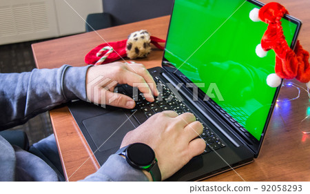 Closeup man hands typing on laptop keyboard with empty green screen and Christmas decoration on background. Holiday concept. Office. Mock-up 92058293