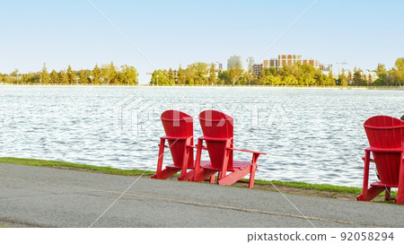 Two Red Adirondack Chairs on the green grass of a pier. Lookout on large blue lake with a perfect blue sky. concept of an ideal resting place. Dow's lake Ottawa, Canada. 92058294