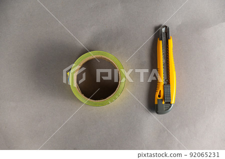 Yellow paper cutter and adhesive tape. The hand tool lies on a gray uneven surface. Blue and yellow duct tape. Top view. Copy space for text and design elements. Yellow paper cutter and adhesive tape. The hand tool lies on a gray uneven surface. Blue and yellow duct tape. Top view. Copy space for text and design elements. 92065231