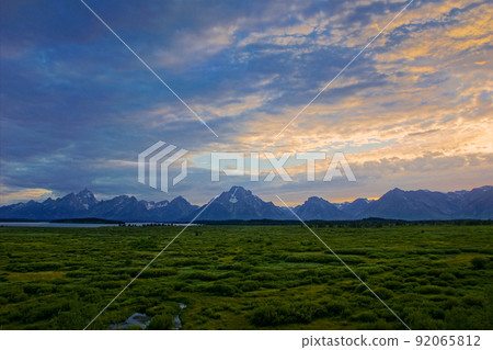 Mountain range of Grand Teton National Park. A distant view of the mountain range and grasslands / lakes with rocky mountains of residual snow that floats dimly at dusk. 92065812