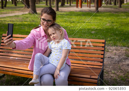 Mother and daughter take a selfie while relaxing on a park bench. 92065893