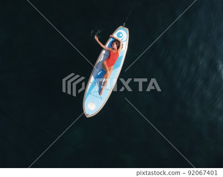 Aerial drone view on well looking middle aged woman with black hair in red swimsuit, swimming on sup in calm azure sea. Summer holiday vacation and travel concept. 92067401