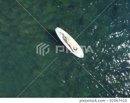 Top view on blonde woman in yellow bikini relaxing on the sup board. Young attractive woman in the sea on the Stand Up Paddle Board SUP. The concept of active and healthy life in harmony with nature. Top view on blonde woman in yellow bikini relaxing on the sup board. Young attractive woman in the sea on the Stand Up Paddle Board SUP. The concept of active and healthy life in harmony with nature. 92067410